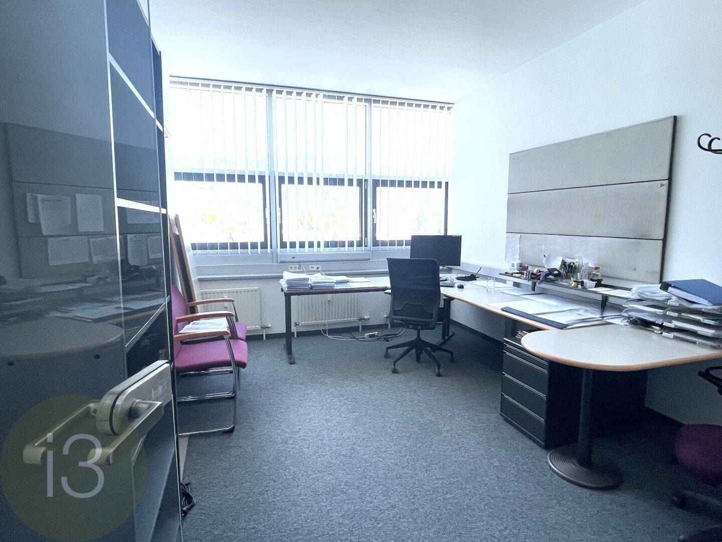 Einzelner Büroraum mit einem Arbeitsplatz, Teppichboden und großen Fenstern mit Jalousien für Privatsphäre.