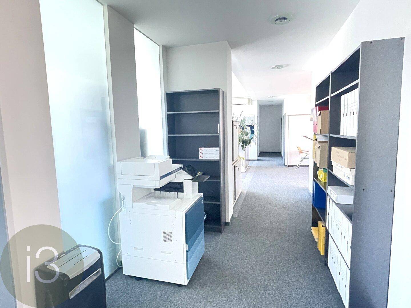 Langer Flur mit Teppichboden, Regalen und einem Kopiergerät, der verschiedene Bürobereiche verbindet.