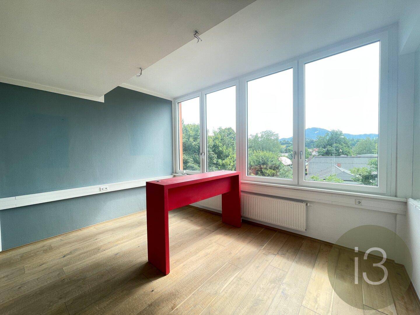 Heller Raum mit blauer Akzentwand, rotem Konsolentisch und großen Fenstern mit Ausblick ins Grüne.