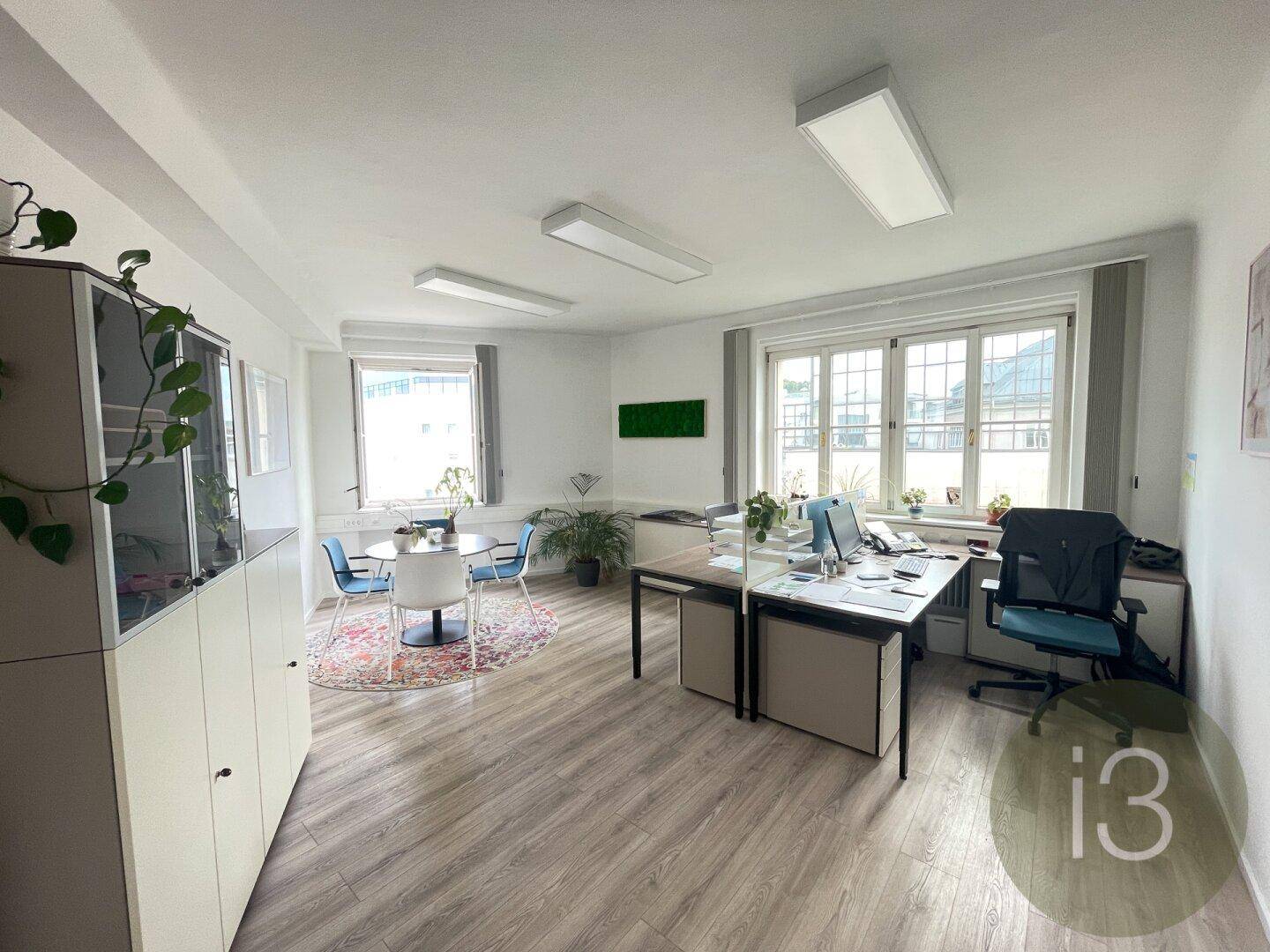 Helles Büro mit mehreren Arbeitsplätzen, Schränken und einem kleinen Besprechungsbereich am Fenster.