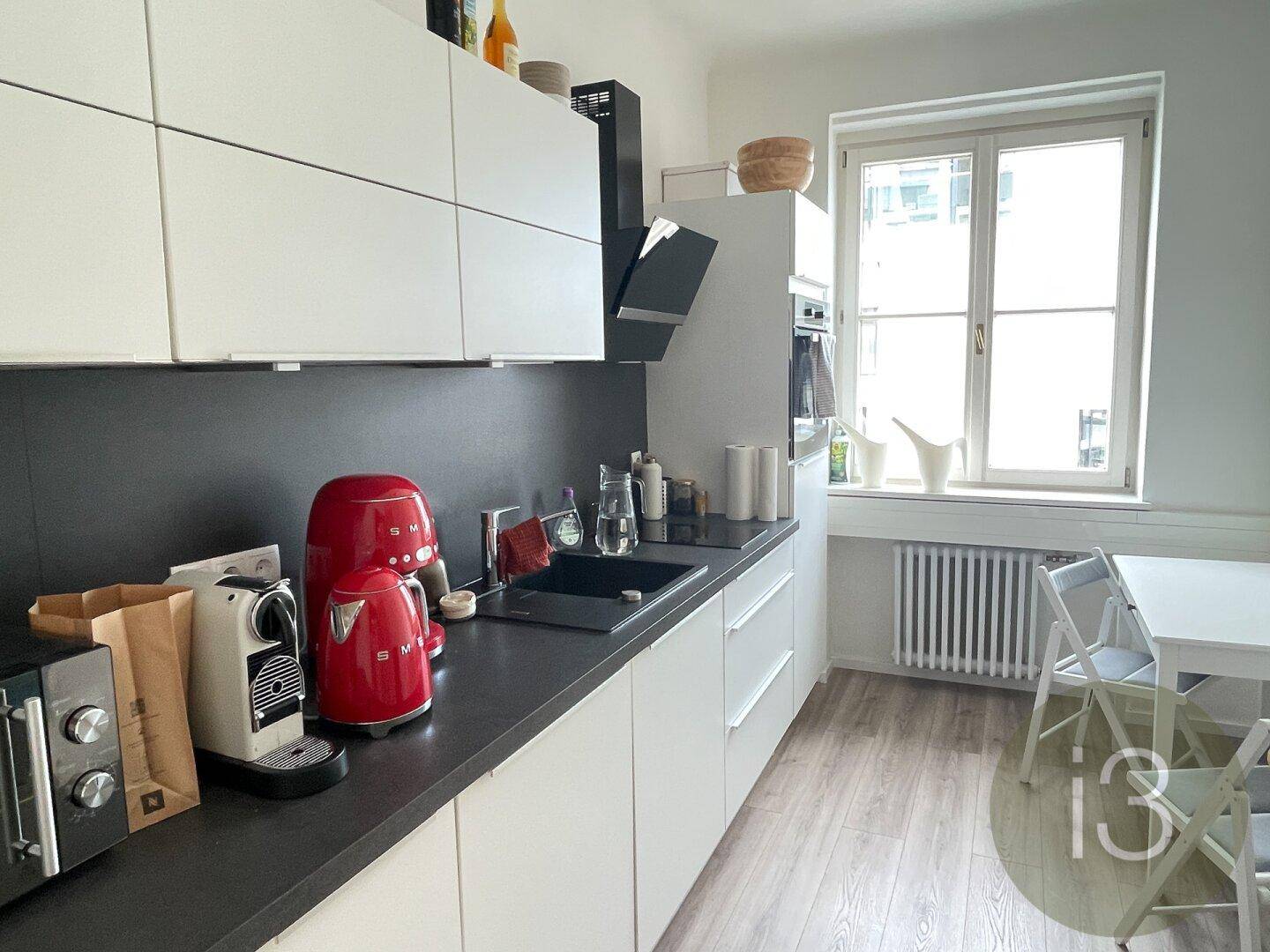 Moderne Küche mit weißen Schränken, dunkler Arbeitsplatte und roten Küchengeräten sowie einem Esstisch.