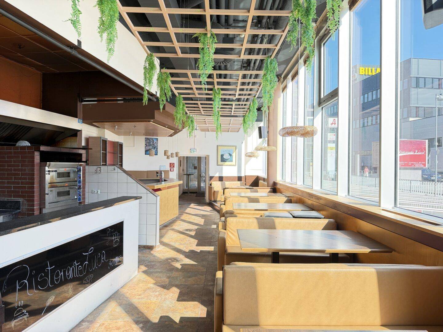 Langer Restaurantraum mit Reihen von Sitzbänken, großen Fenstern und einer sichtbaren Pizzaofen-Küche im Hintergrund.