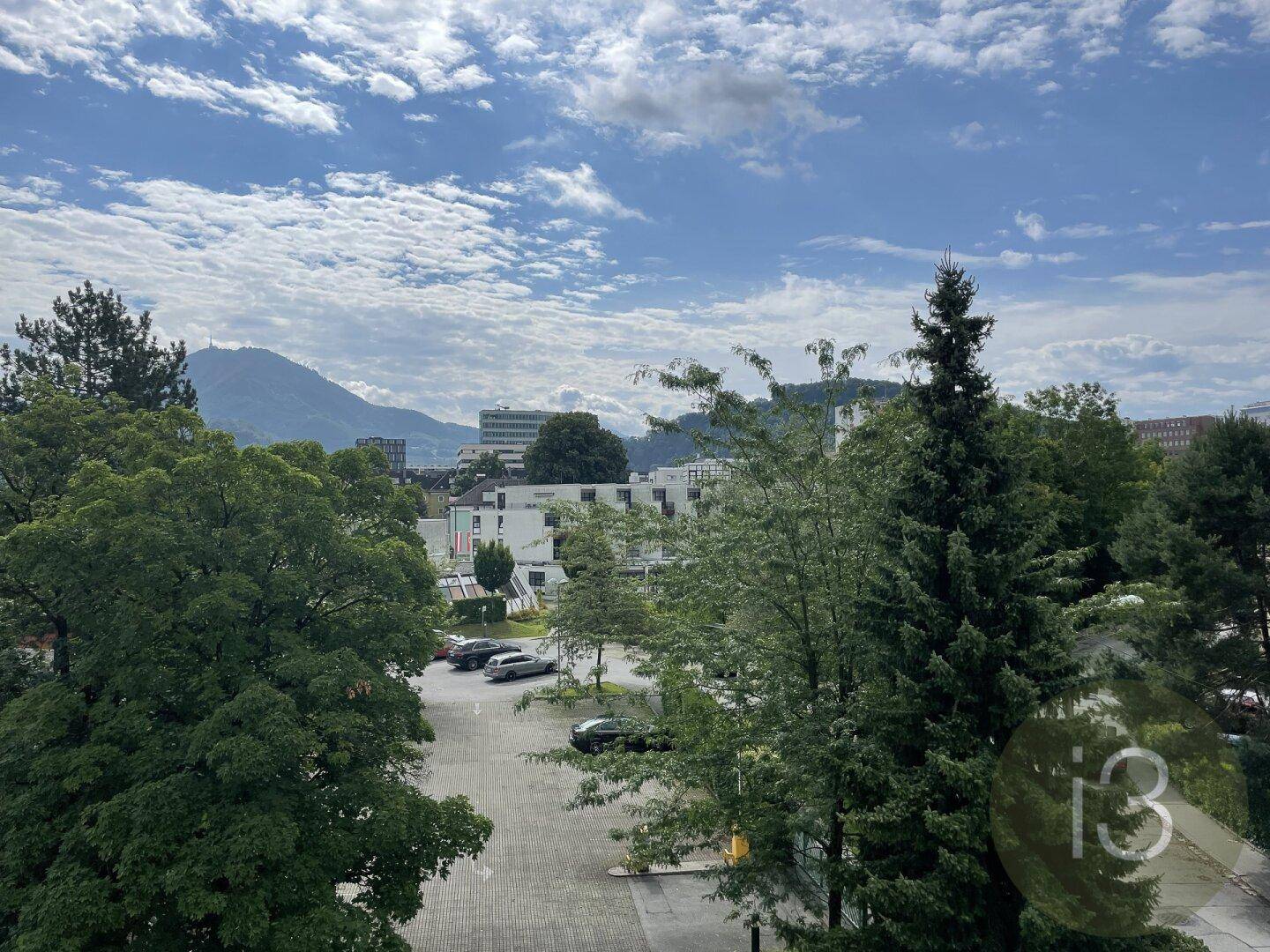 Weitläufiger Ausblick über grüne Bäume, einen Parkplatz und die umliegende Berglandschaft.
