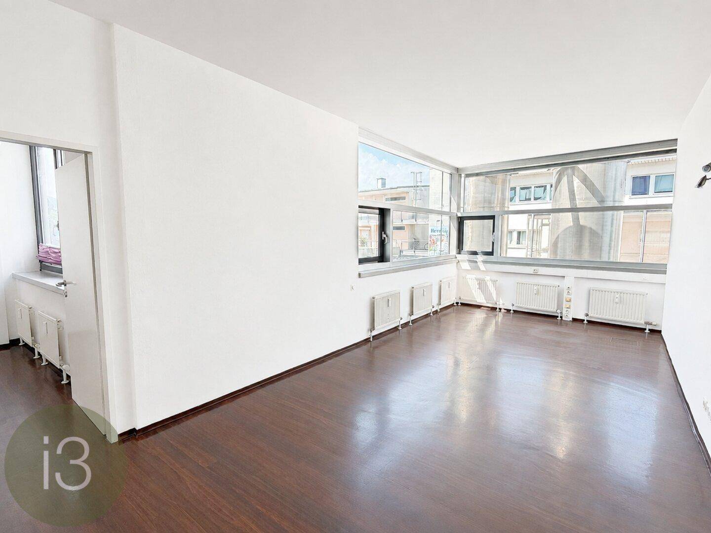 Heller Raum mit großen Fenstern, dunklem Holzboden und Blick auf umliegende Gebäude.