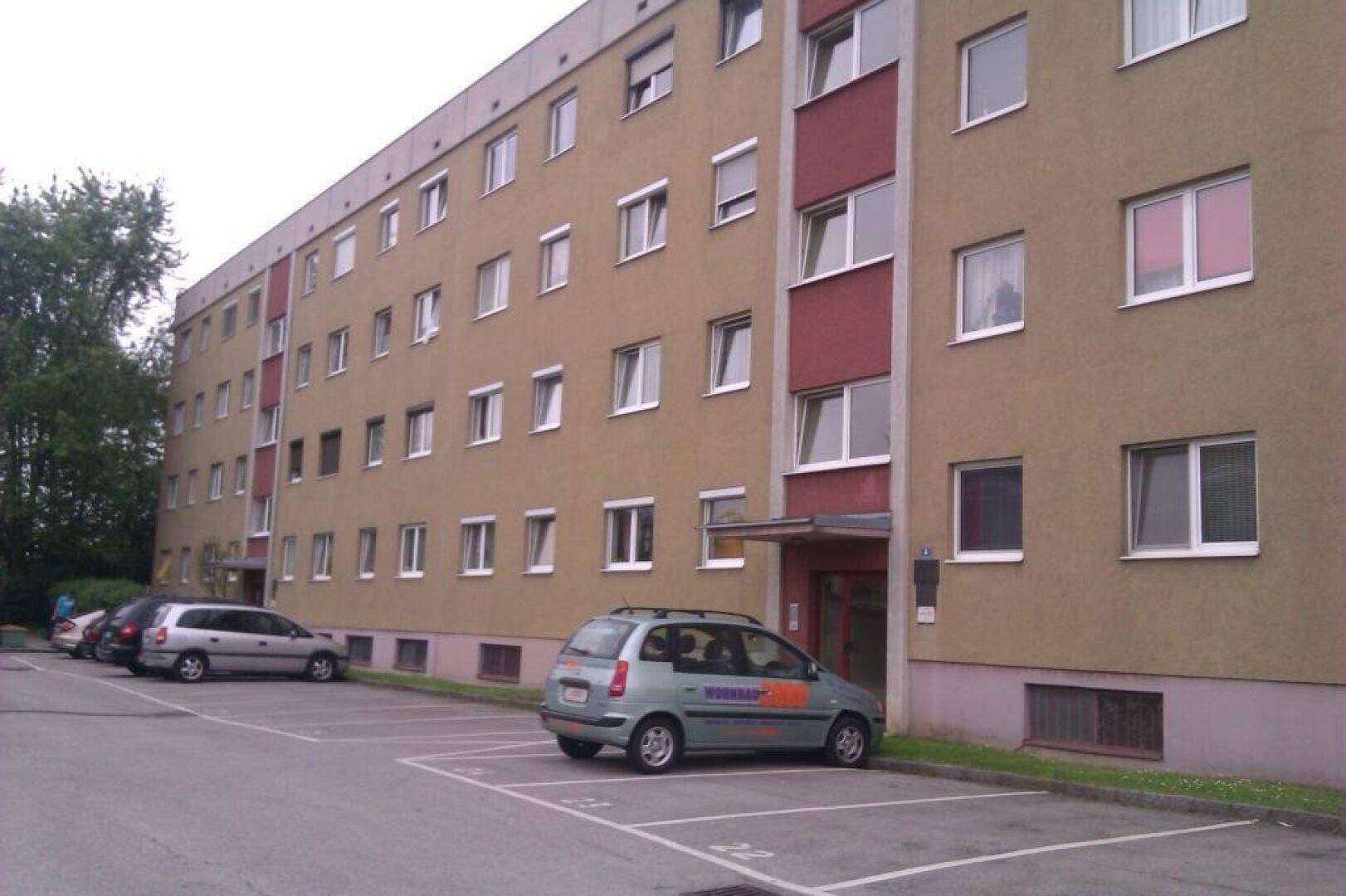 Langes Wohngebäude mit beiger und roter Fassade, umgeben von Parkplätzen und Bäumen.