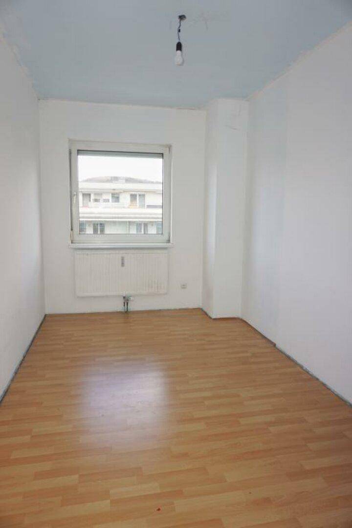 Helles, leeres Zimmer mit Laminatboden, Fenster und einer hellblauen Decke.