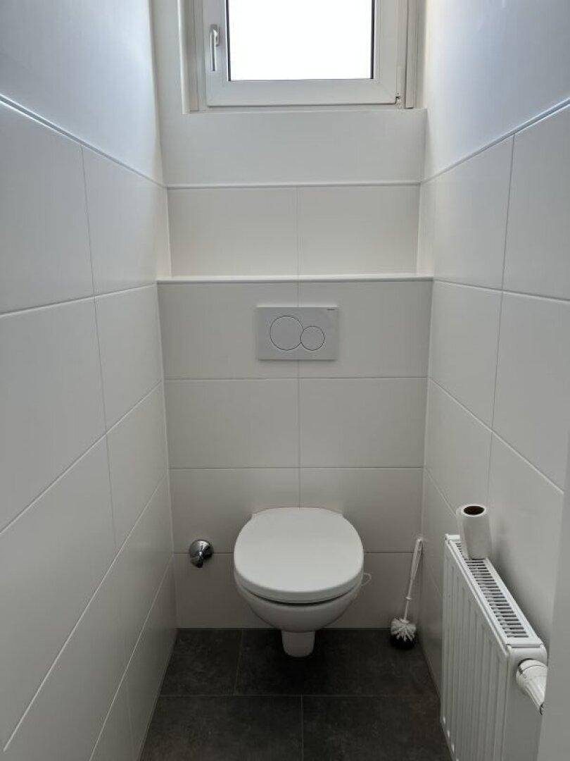 Kleines Gäste-WC mit wandhängender Toilette, Fenster und weißen Wandfliesen.