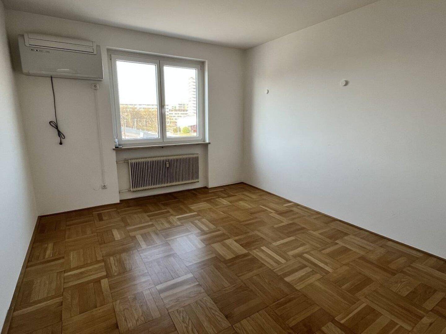 Einfaches Zimmer mit Parkettboden, einem Fenster und einer Klimaanlage an der Wand.