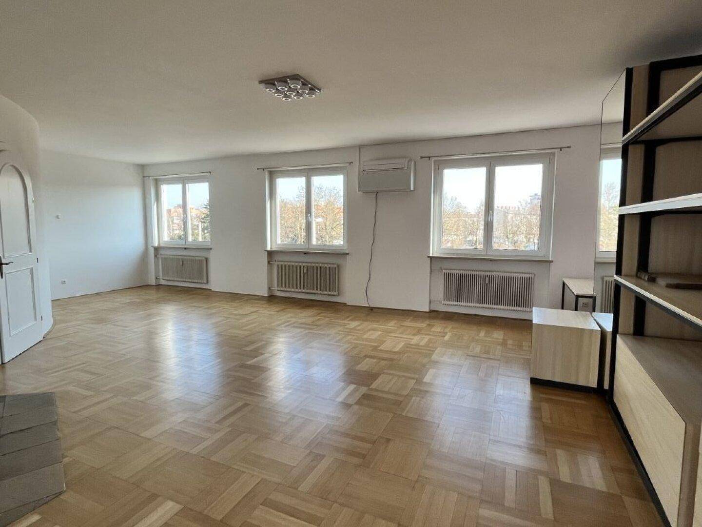 Großes, helles Wohnzimmer mit Parkettboden, mehreren Fenstern und einer Klimaanlage.