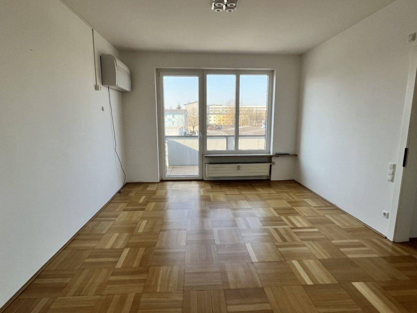 Helles Zimmer mit Parkettboden, großem Fenster und Zugang zu einem möglichen Balkon.