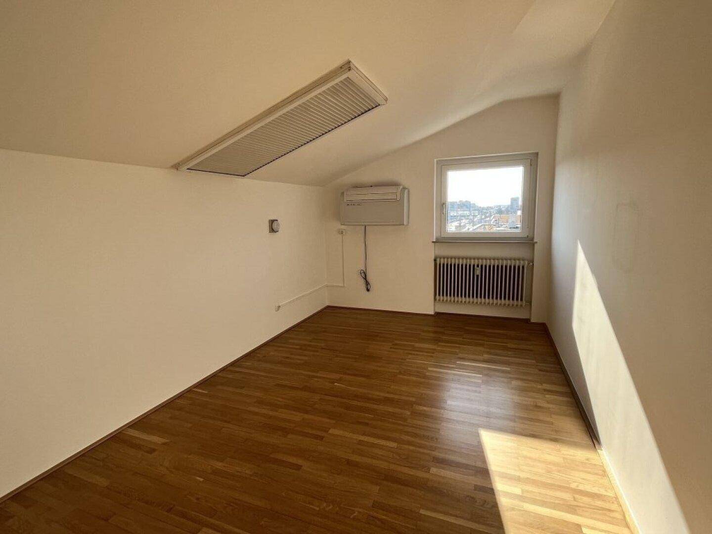 Helles Dachzimmer mit Parkettboden, schräger Decke und einem Fenster mit Stadtblick.