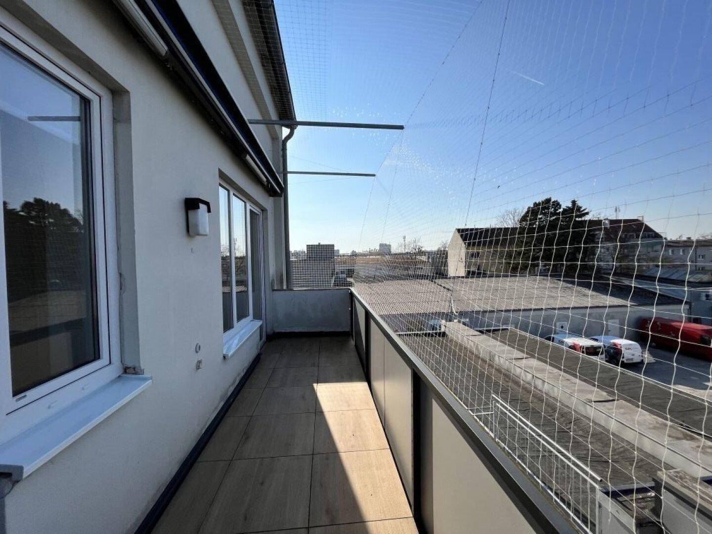 Langer Balkon mit Schutznetz, Fliesenboden und Blick auf die städtische Umgebung.