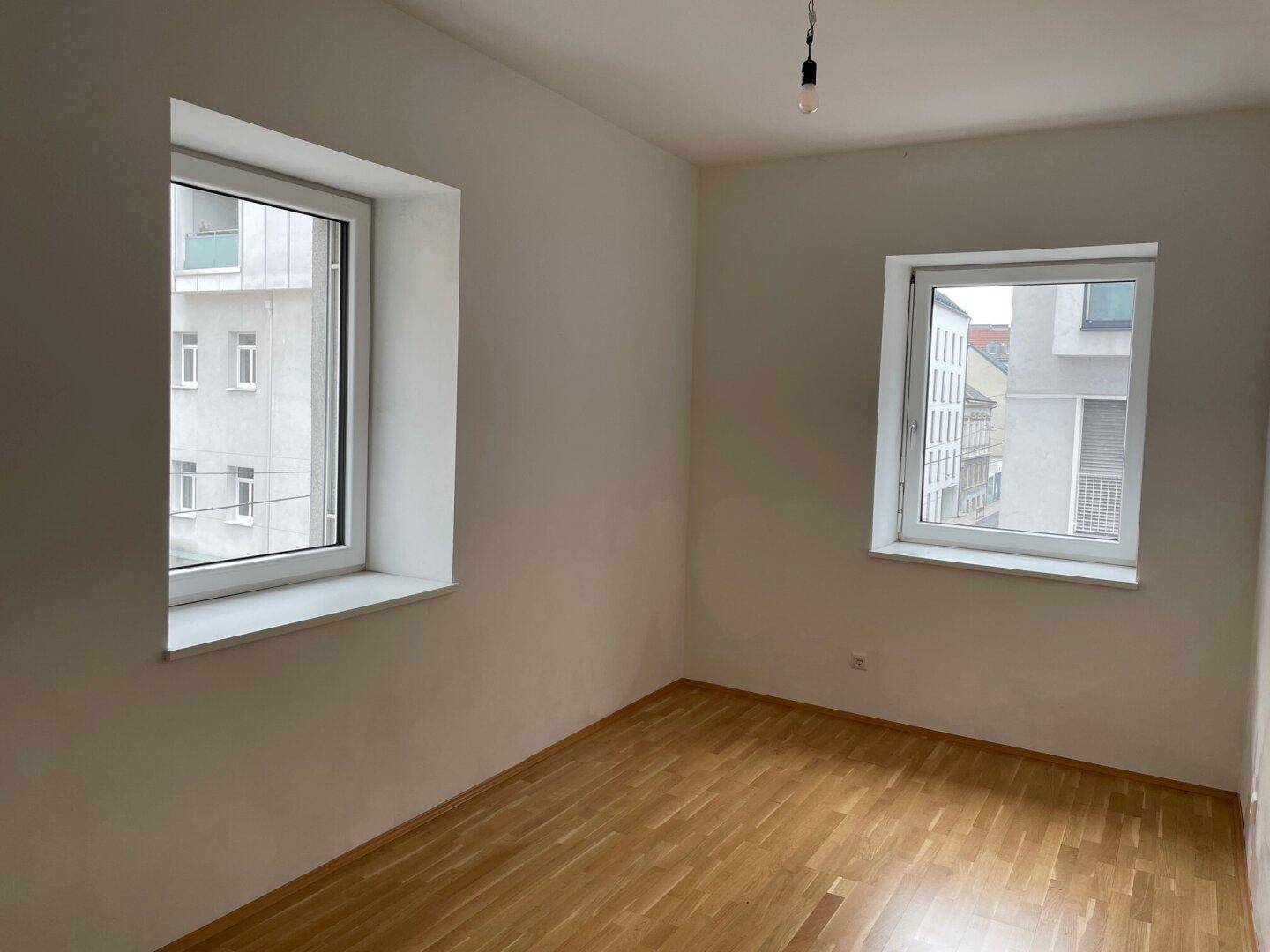 Freundliches Zimmer mit zwei Fenstern und Holzboden, das viel Tageslicht hereinlässt.