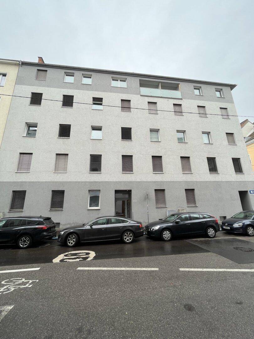 Mehrstöckiges Wohngebäude mit grauer Fassade, vielen Fenstern und Jalousien an einer Straße.