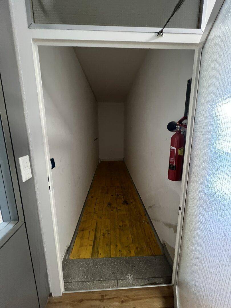 Schmaler Abstellraum oder Lagerbereich mit Holzboden und einem Feuerlöscher an der Wand.