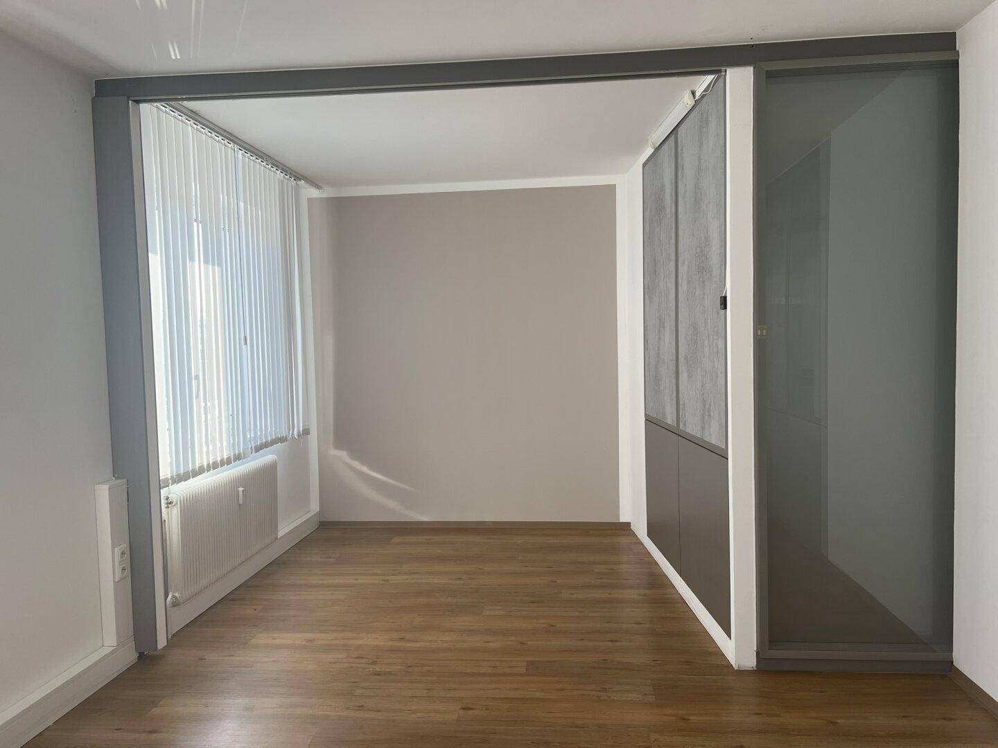 Heller Büroraum mit Holzboden, hellgrauen Wänden und großen Fenstern mit Jalousien.