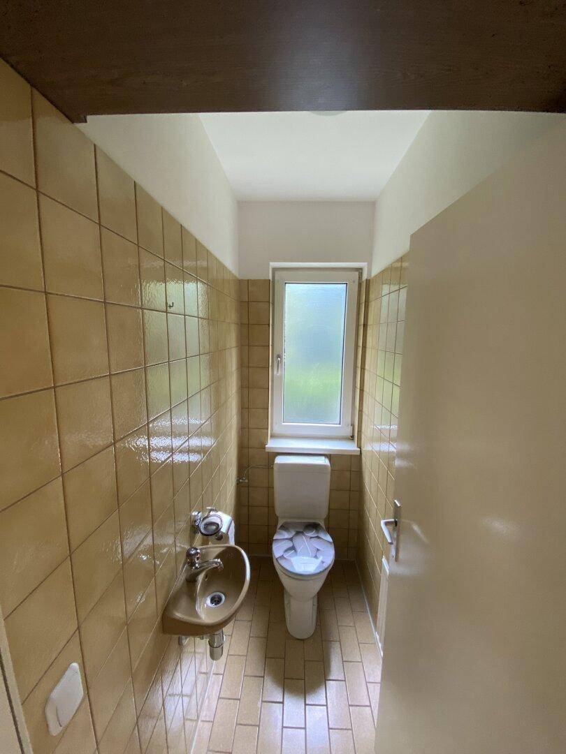 Gäste-WC mit beigefarbenen Fliesen, Toilette, kleinem Waschbecken und Fenster.