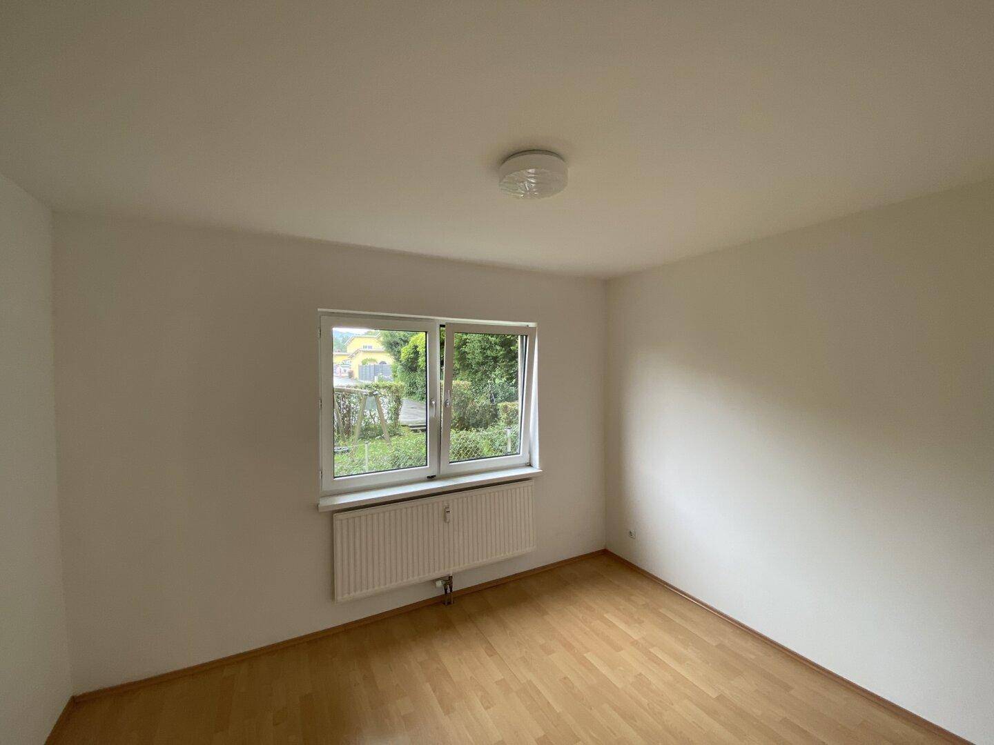 Leeres Schlafzimmer mit Laminatboden, weiß gestrichenen Wänden und Blick ins Grüne.
