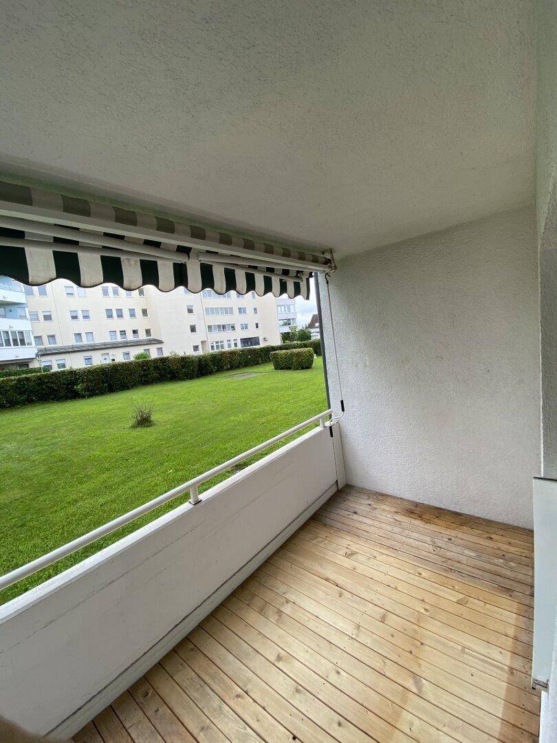 Balkon mit Holzboden, Markise und Blick auf eine grüne Rasenfläche und umliegende Gebäude.