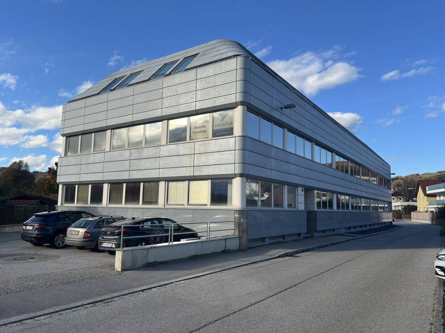 Modernes Bürogebäude mit markanter Fassade, großen Fenstern und ausreichend Parkplätzen im Freien.
