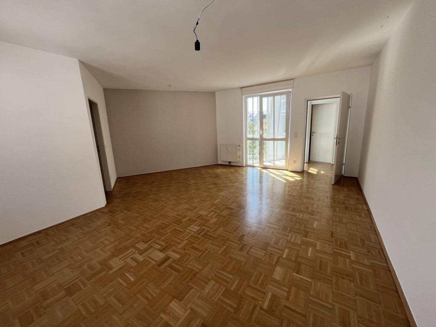 Helles leeres Zimmer mit Parkettboden und großem Fenster, das viel Tageslicht hereinlässt.