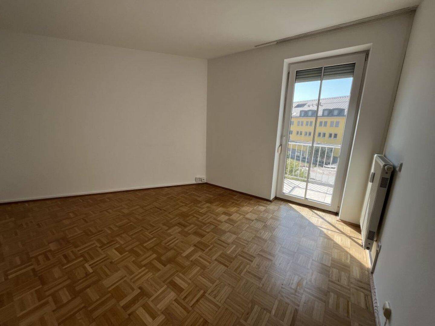 Helles leeres Zimmer mit Parkettboden und Zugang zu einem französischen Balkon.