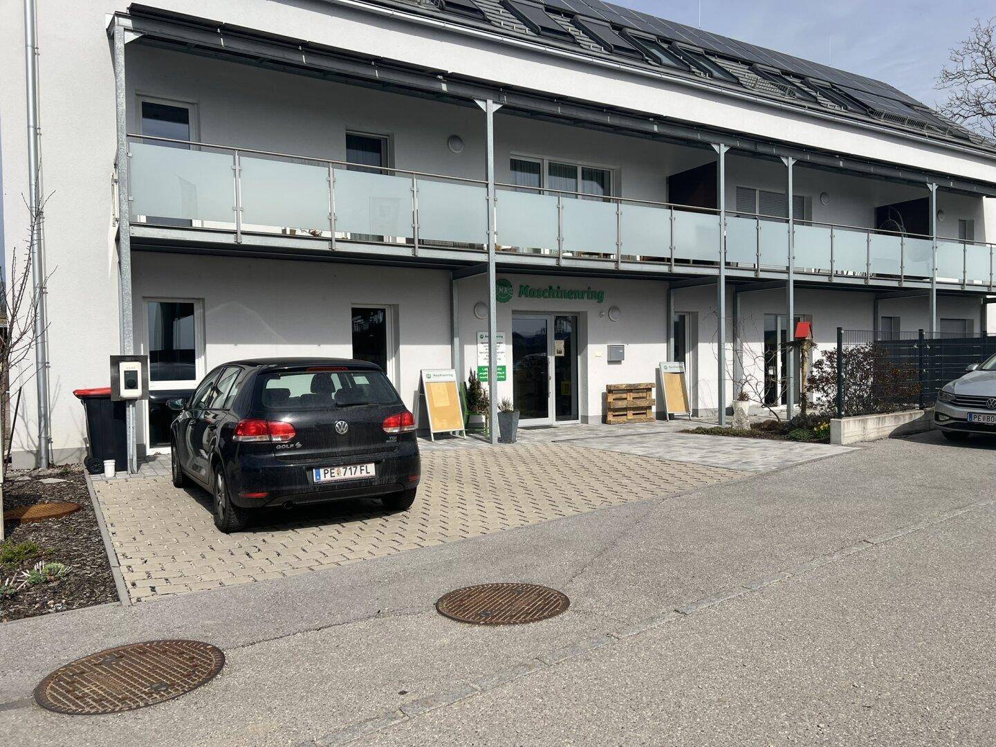 Modernes Geschäftsgebäude mit Solarpaneelen auf dem Dach und Parkplätzen davor, umgeben von Bäumen.