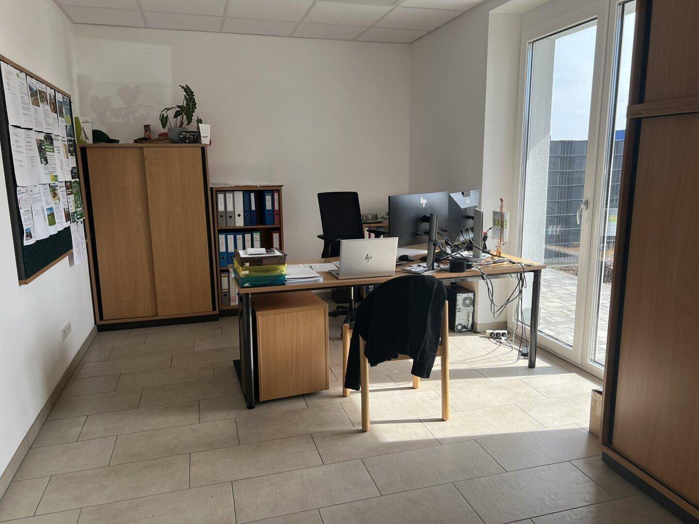 Geräumiges Büro mit großem Schreibtisch, Holzschränken und viel Tageslicht durch die bodentiefen Fenster.