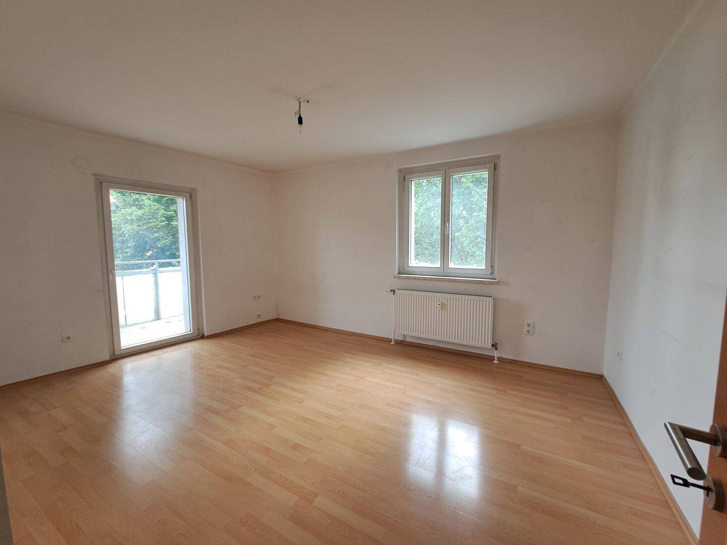 Helles, leeres Wohnzimmer mit Laminatboden, Fenster und Zugang zu einem Balkon.