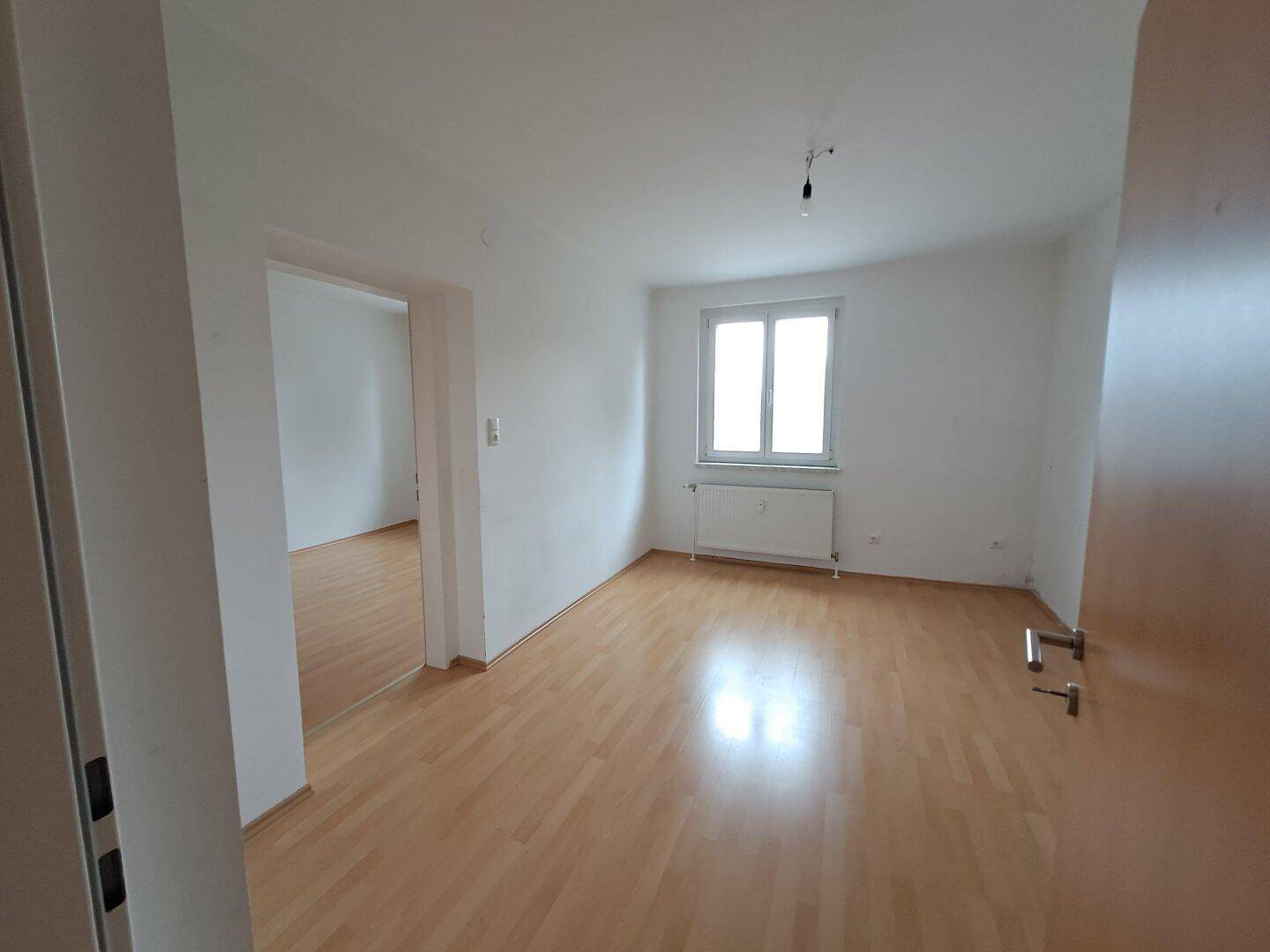 Geräumiges, leeres Zimmer mit Laminatboden und weiß gestrichenen Wänden, offen zu einem weiteren Raum.