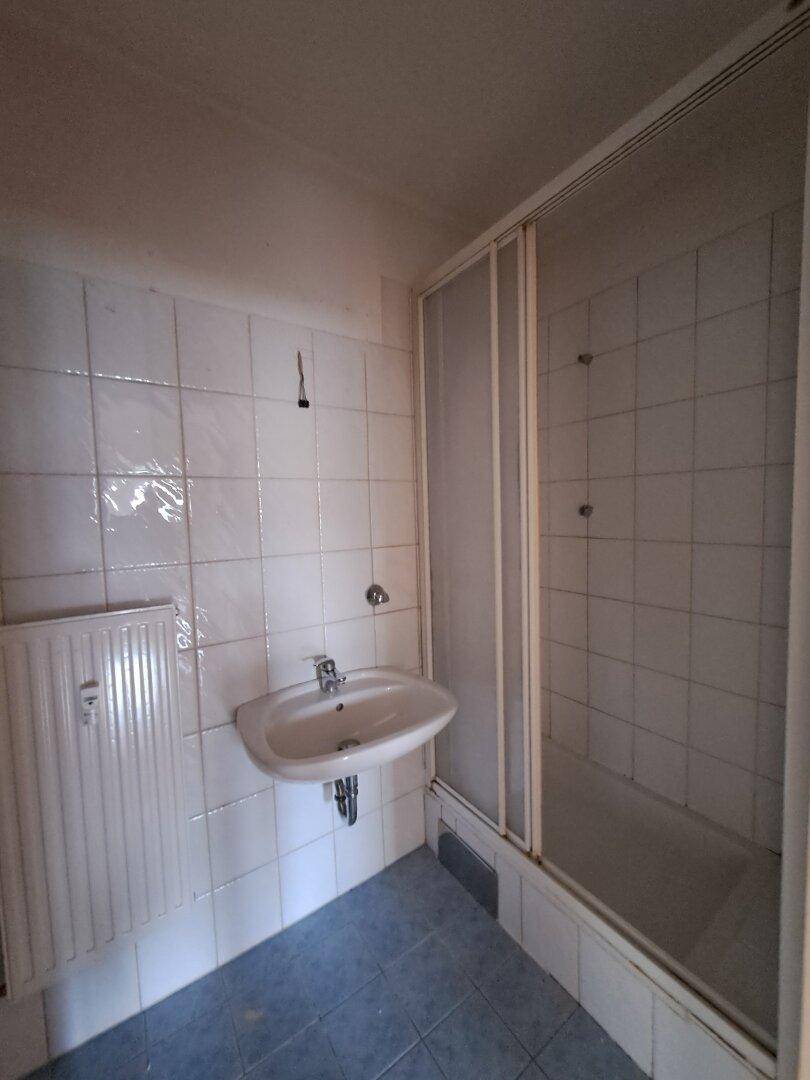Badezimmer mit weißen Fliesen, Waschbecken und einer Duschkabine, in älterem Zustand.