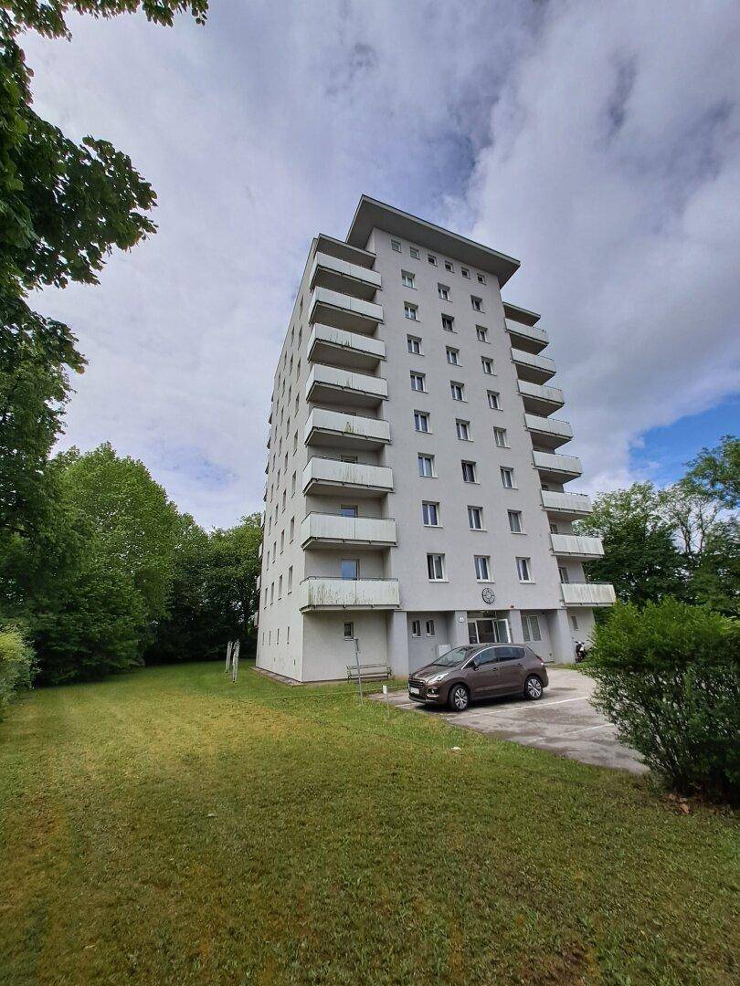 Hohes Wohngebäude mit vielen Balkonen und umliegender Grünfläche unter bewölktem Himmel.