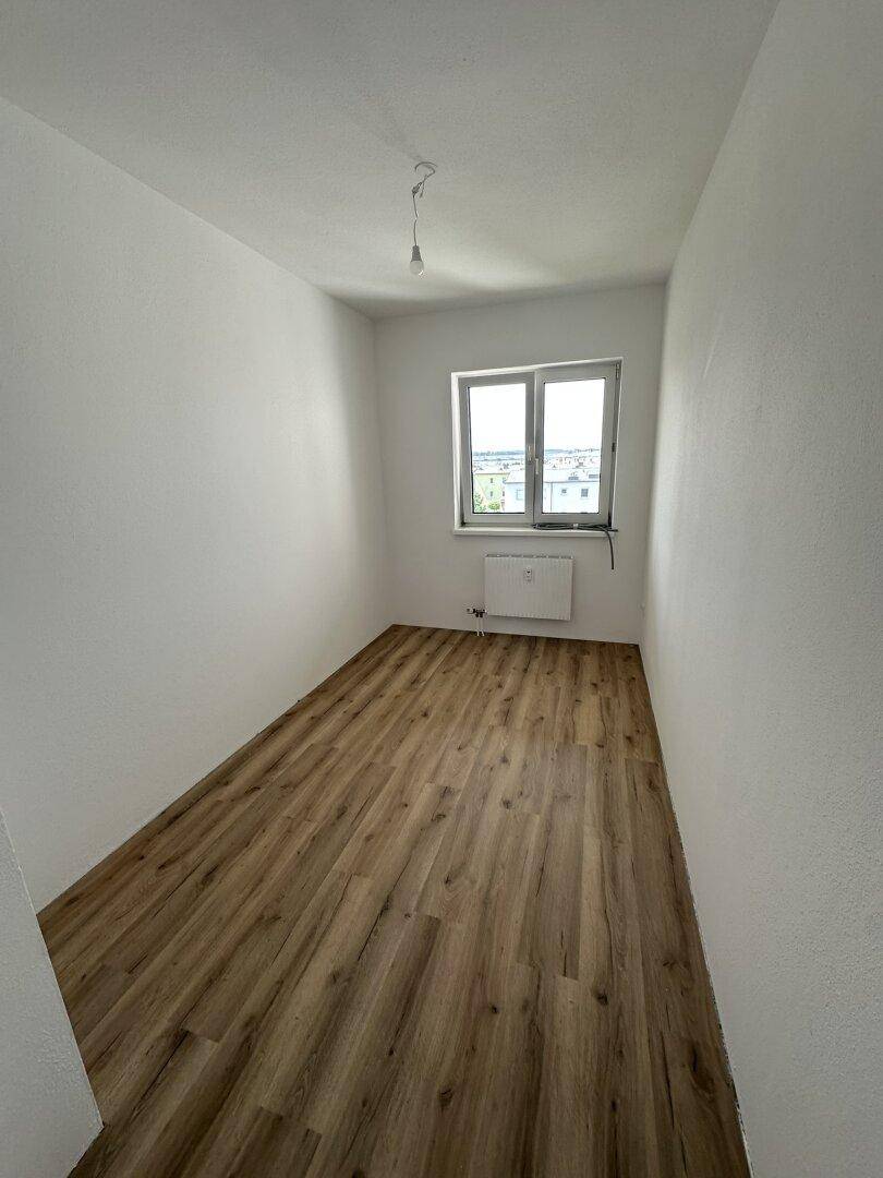 Schmaler, heller Raum mit Laminatboden, weißen Wänden und einem Fenster für natürliche Belichtung.