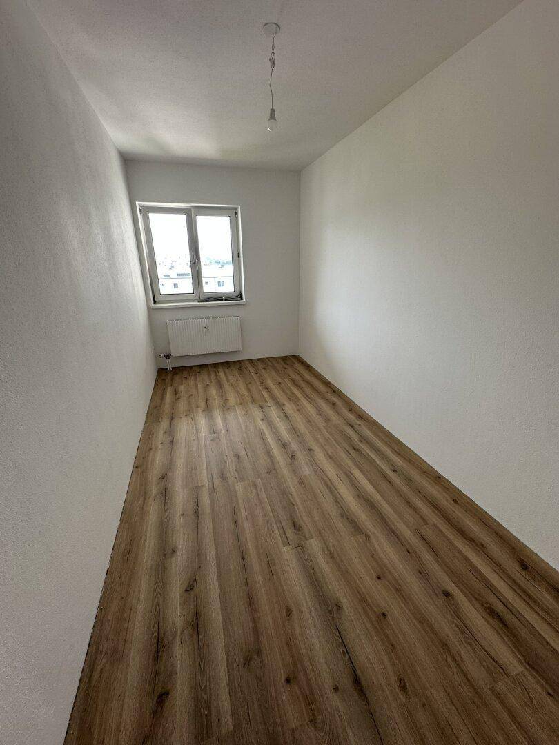 Heller, leerer Raum mit Laminatboden und einem Fenster, das viel Tageslicht hereinlässt.