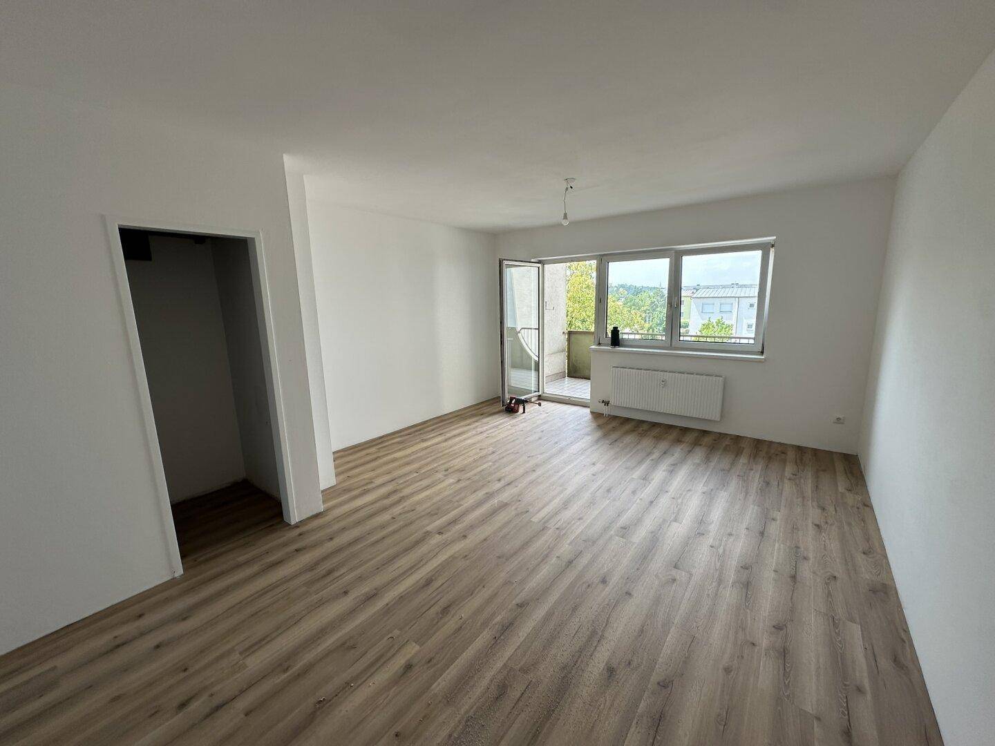 Helles, leeres Zimmer mit neuem Laminatboden, weißen Wänden und Zugang zu einem Balkon.