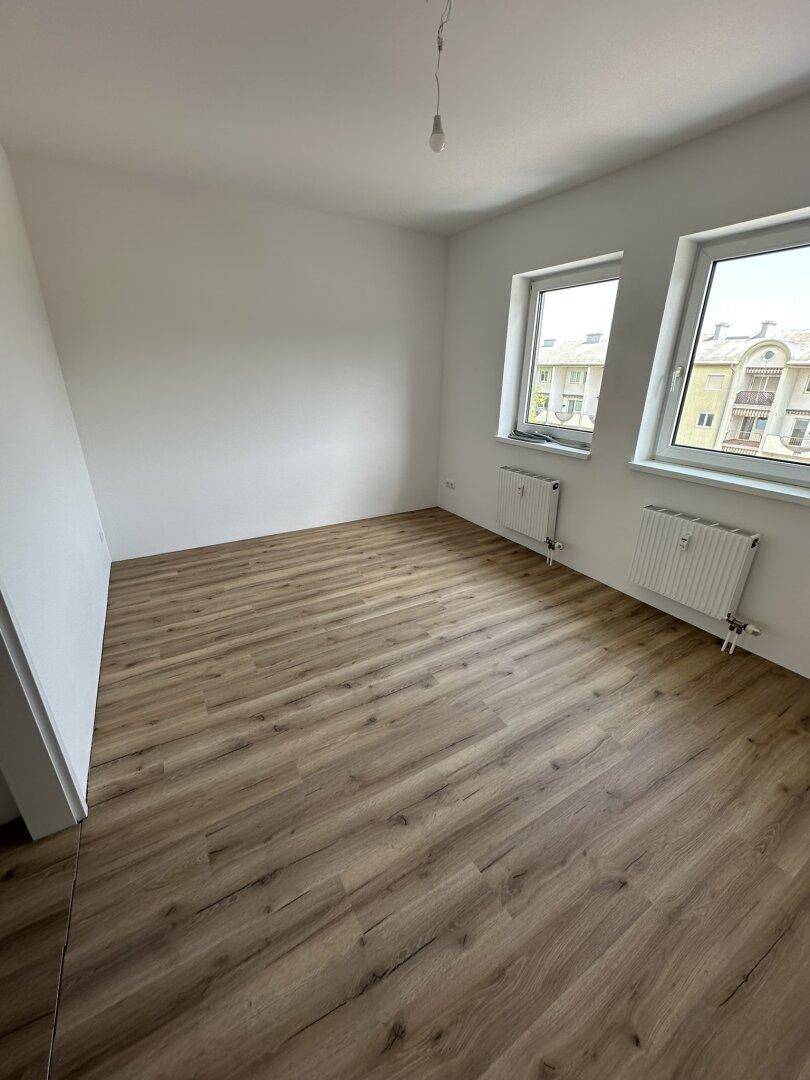 Leeres Zimmer mit hellem Laminatboden, weißen Wänden und zwei Fenstern für viel Tageslicht.