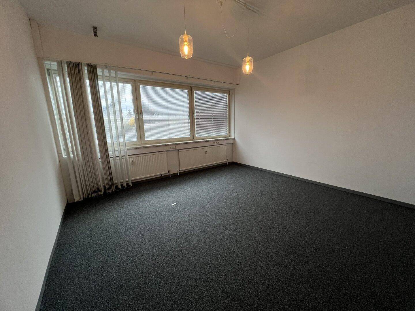 Helles Büro mit Teppichboden, großen Fenstern und modernen Hängelampen, ideal für Arbeitsbereiche.