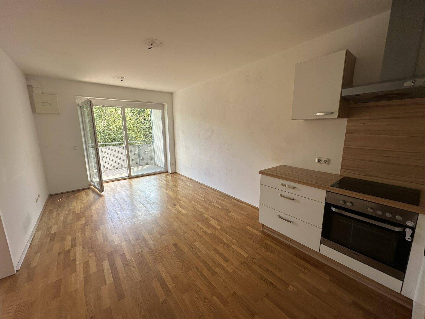 Heller offener Wohnbereich mit integrierter Küchenzeile, Holzboden und Zugang zum Balkon.
