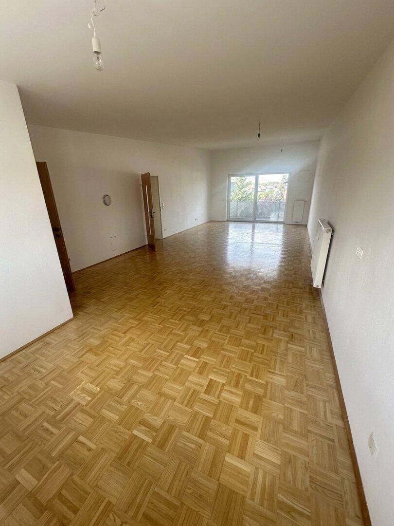 Heller, leerer Wohnbereich mit Parkettboden und Zugang zu einem Balkon oder einer Terrasse.