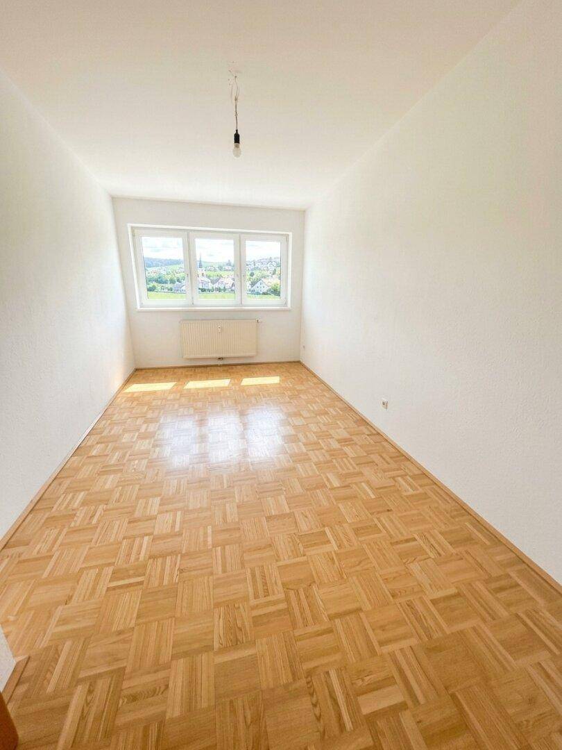 Helles Schlafzimmer mit Parkettboden und drei Fenstern, die einen weiten Ausblick bieten.