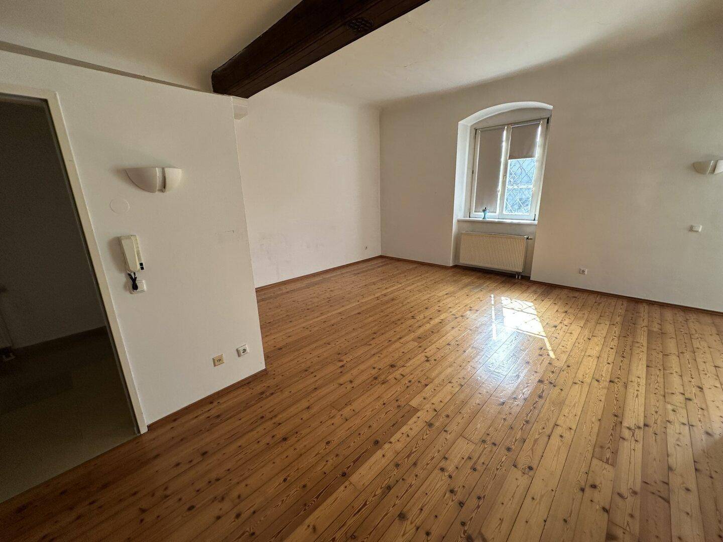 Heller Wohnraum mit Holzdielenboden, sichtbarem Deckenbalken und einem Fenster mit Heizkörper.
