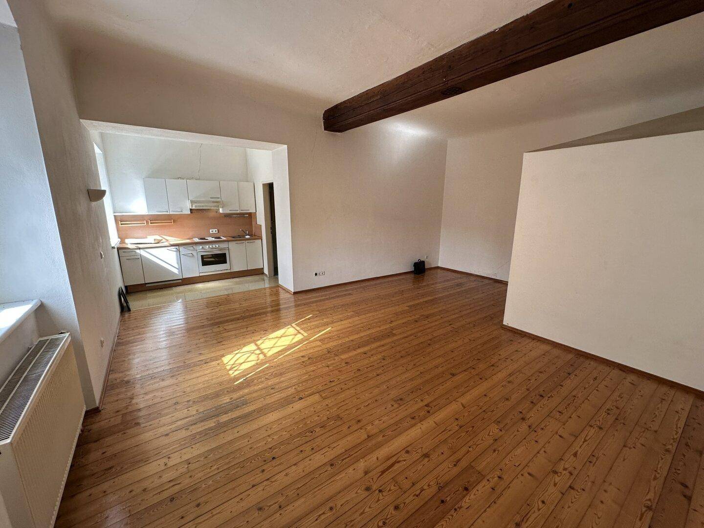 Heller Wohnbereich mit Holzdielenboden, sichtbarem Deckenbalken und integrierter Küchenzeile.