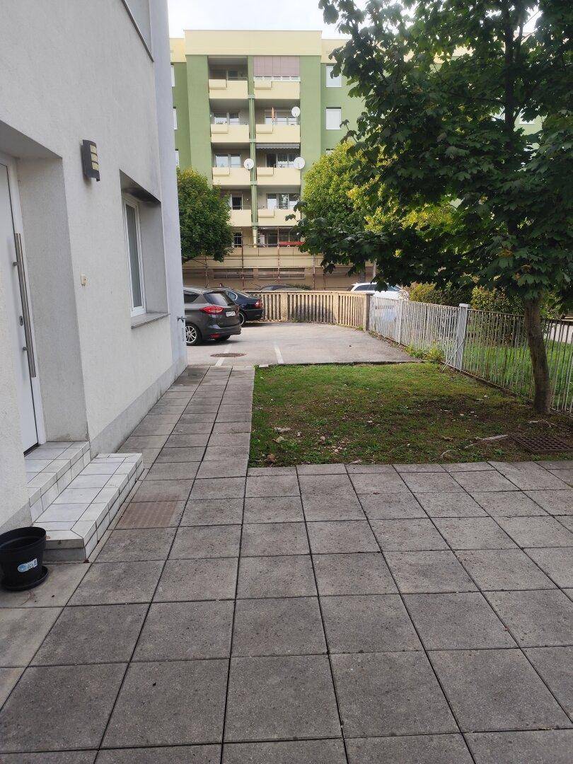 Seitenansicht des Gebäudes mit gepflastertem Bereich, kleiner Rasenfläche und Parkplätzen im Hintergrund.
