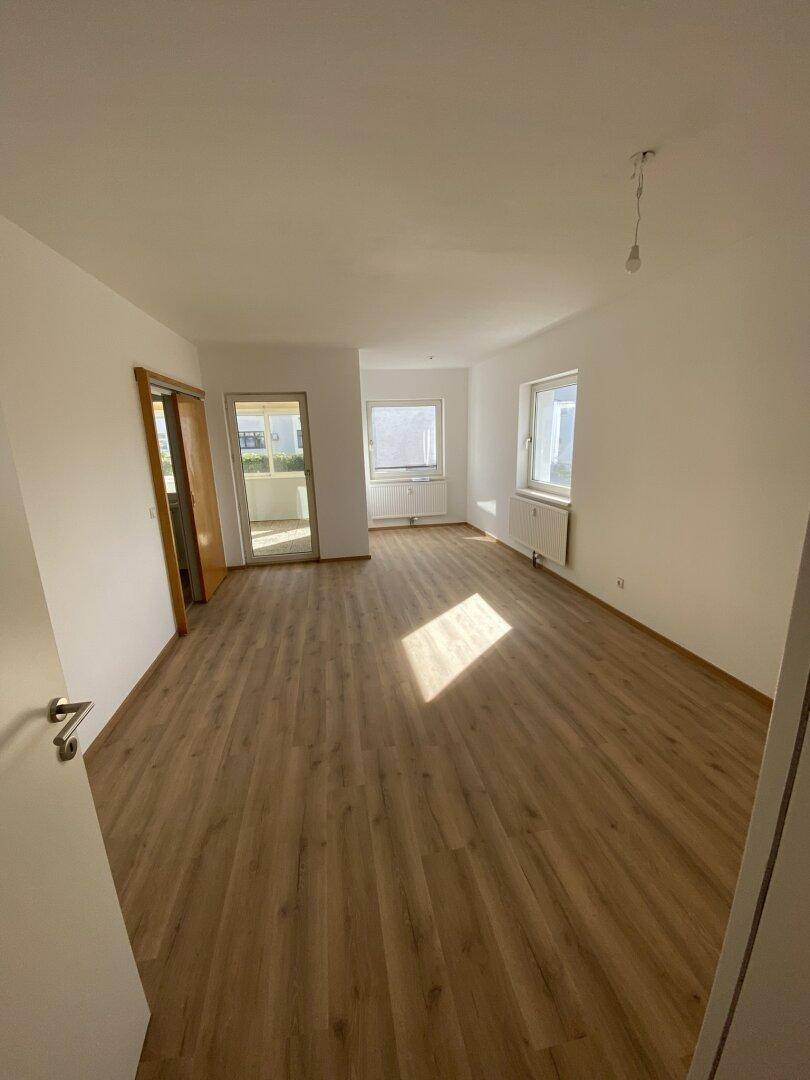 Heller, leerer Wohnbereich mit Laminatboden und Zugang zu einem Balkon.