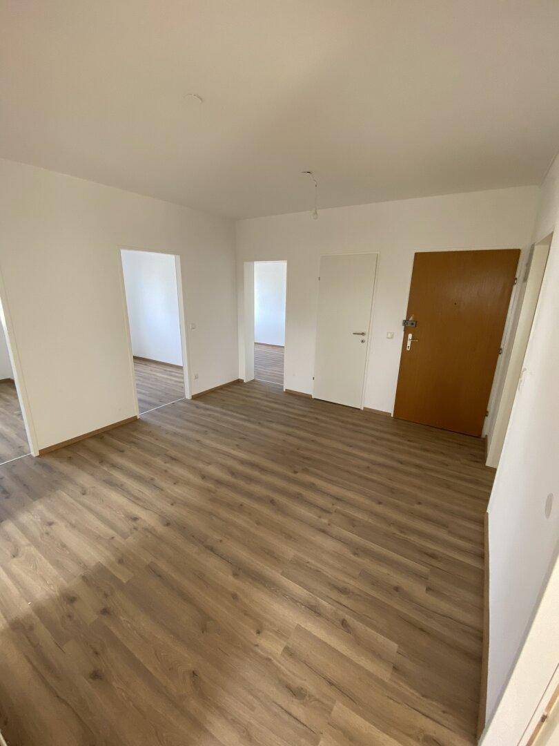 Leerer Wohnraum mit Laminatboden und Zugang zu weiteren Zimmern und der Eingangstür.