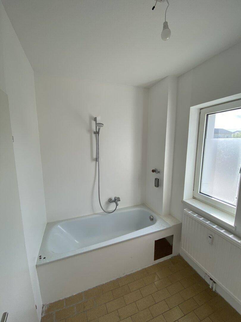 Helles Badezimmer mit Badewanne, Duscharmatur und Fenster, bereit zur Modernisierung.