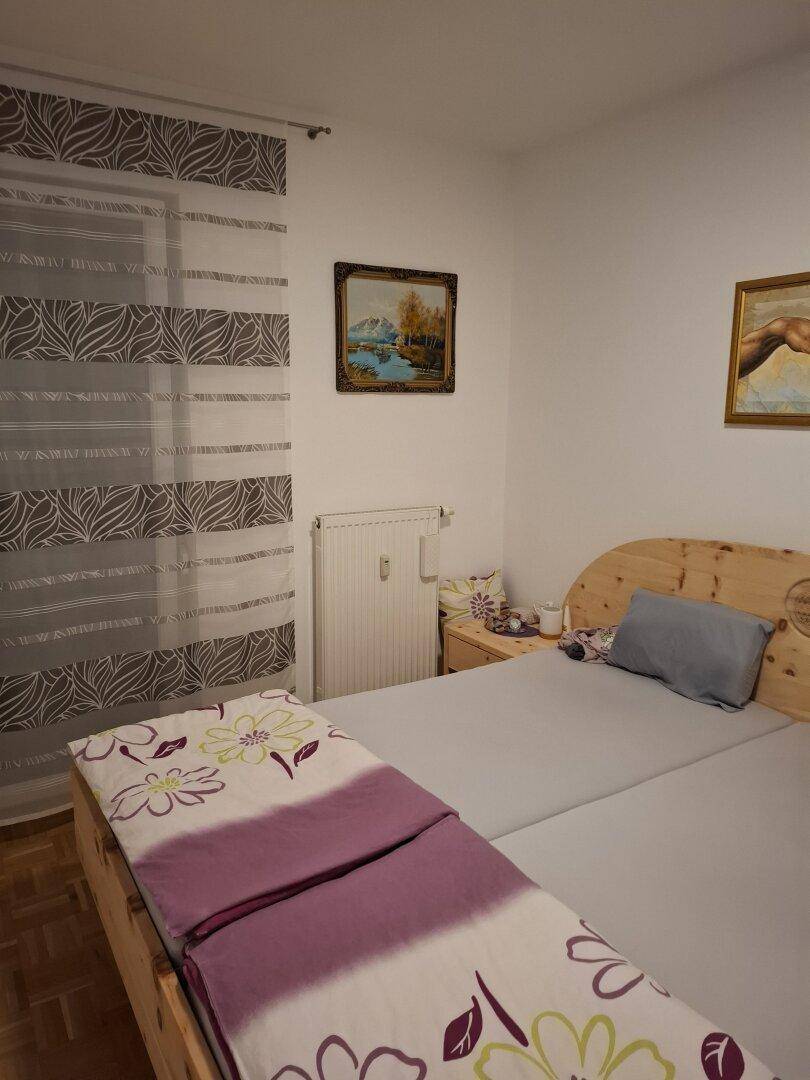 Schlafzimmer mit hellem Holzbett, Nachttisch und Fenster mit gemusterten Vorhängen.
