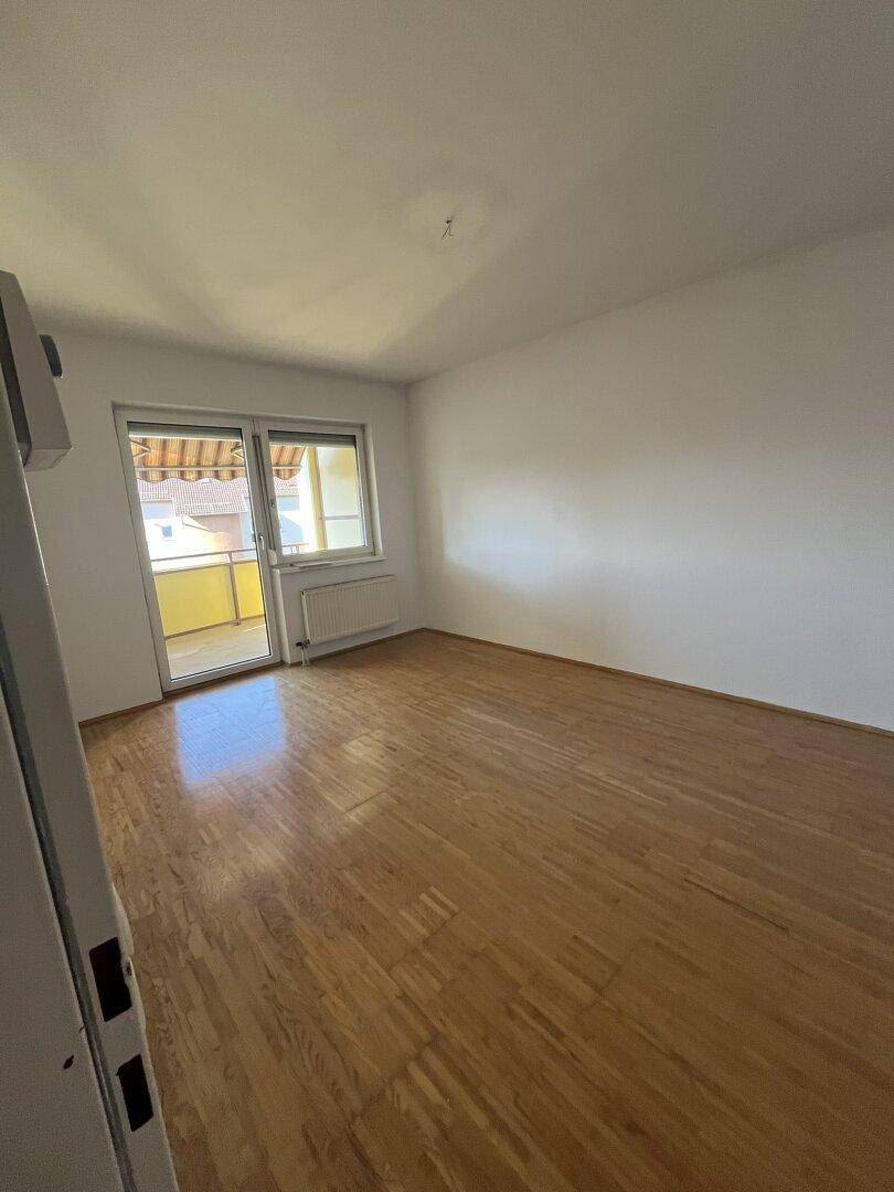 Heller, leerer Raum mit glänzendem Parkettboden, weißen Wänden und Zugang zu einem sonnigen Balkon.