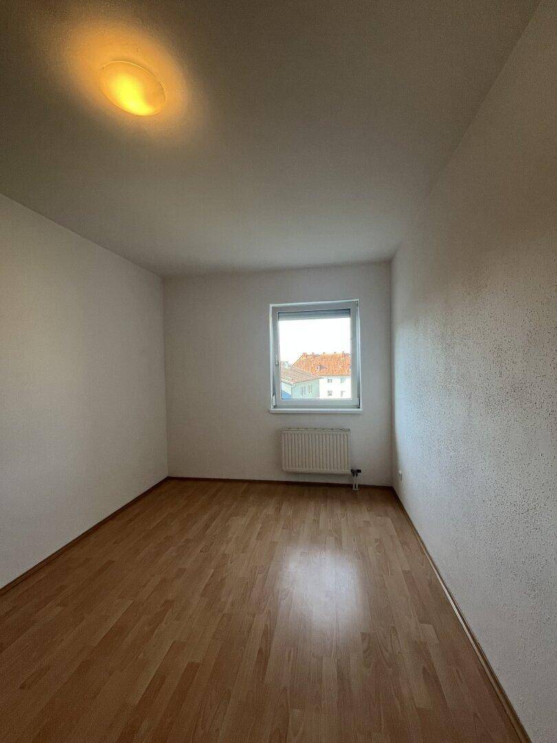 Heller, leerer Raum mit Parkettboden, weißen Wänden und einem Fenster mit Heizkörper darunter.