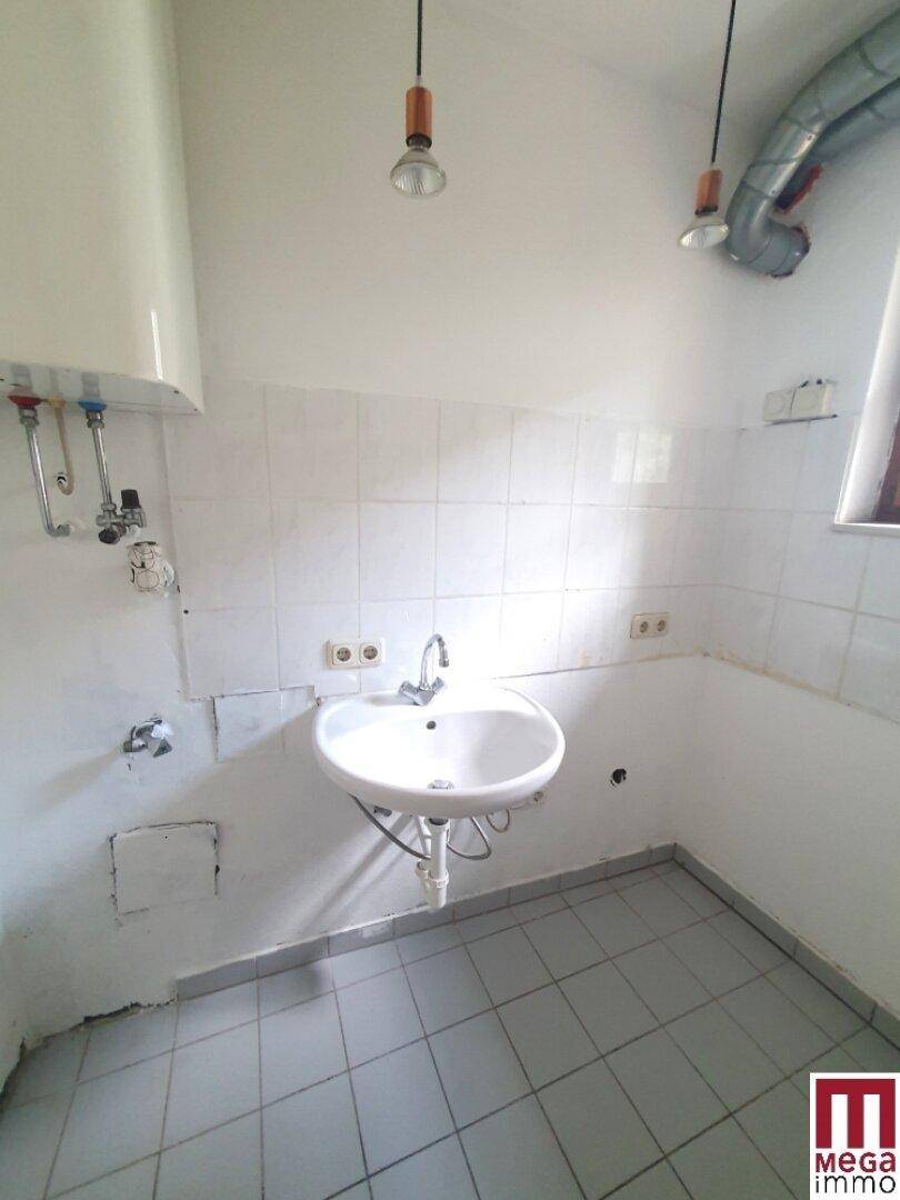 Kleiner Raum mit Waschbecken, weißen Fliesen und sichtbaren Leitungen, möglicherweise für eine Teeküche oder Reinigung.